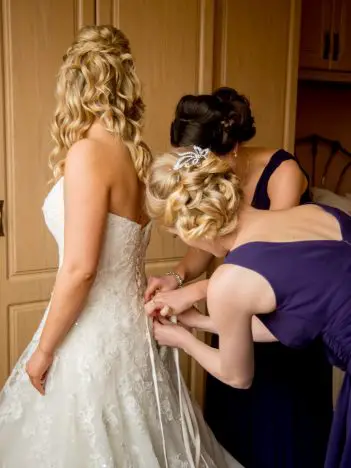 A bride in a corset wedding dress that is being done up by 2 bridesmaids in purple dresses
