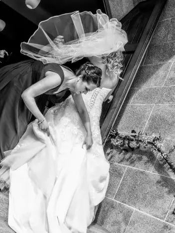 A black and white image of a bride arriving at the church with her hair blowing in the wind. A bridesmaid is helping pull her dress up the stairs