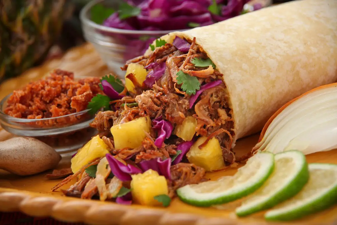 A pulled beef burrito with pineapple chunks, laid on a large board with lime wedges and condiments