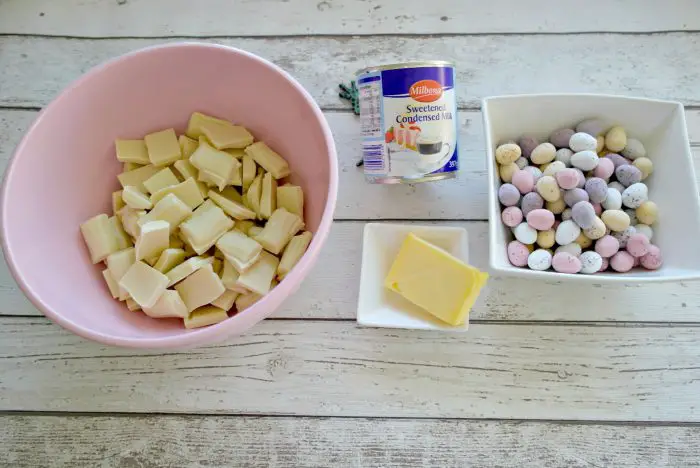 A pink bowl containing broken up white chocolate, a white dish containing a slab of butter, a tin of condensed milk and a white bowl of mini eggs - the ingredients for Easter fudge