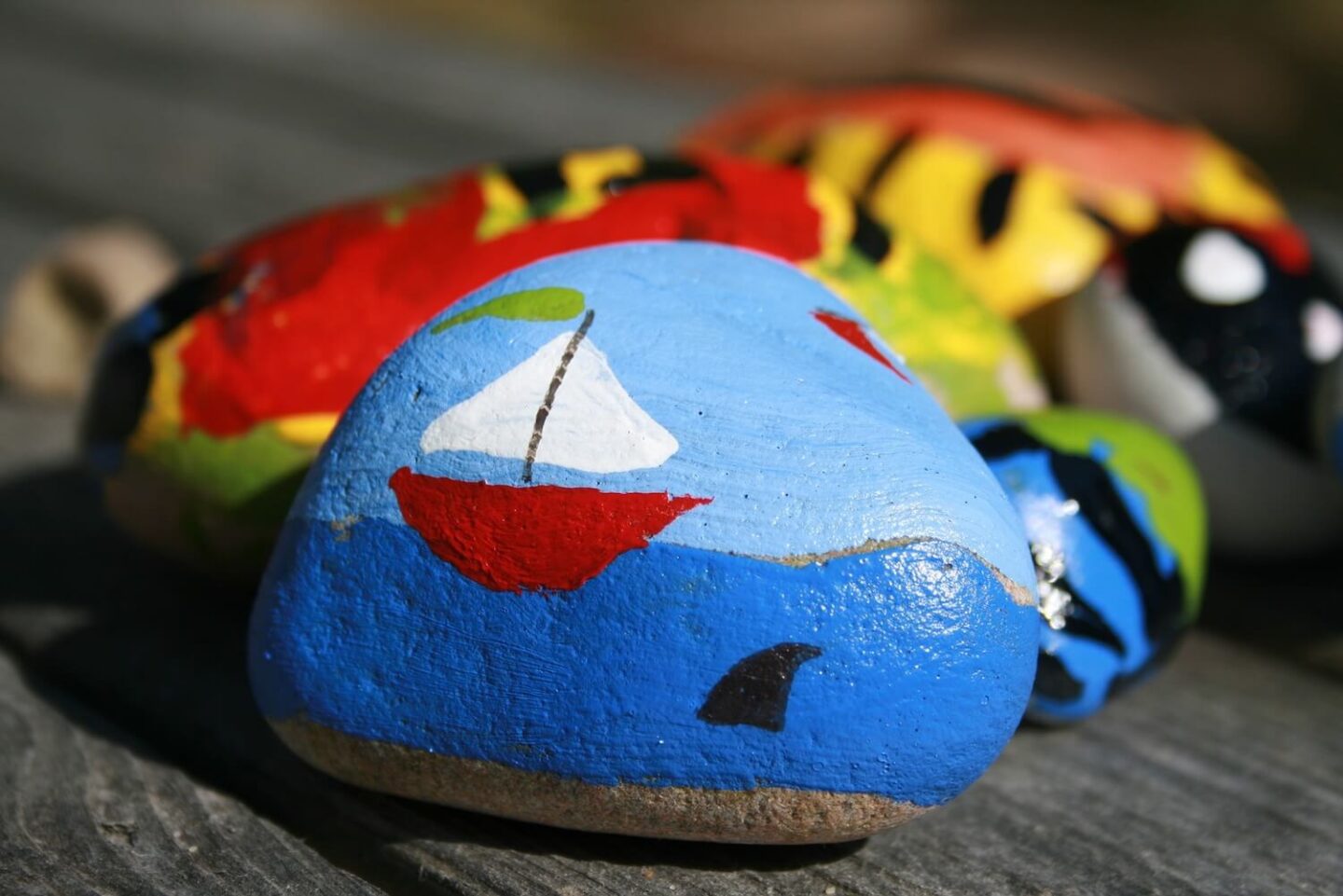 A pile of painted pebbles. The one closest to the camera depicts a sail boat on the ocean