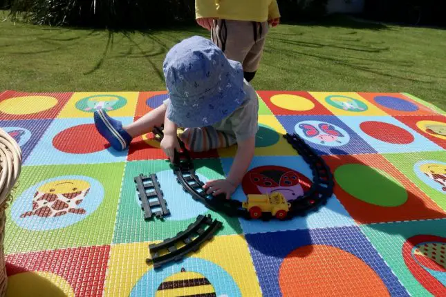 A young boy lying on a cushioned playmat, playing with a train track. The mat has a pattern made up of circles and squares in bright colour, with some zoo animals