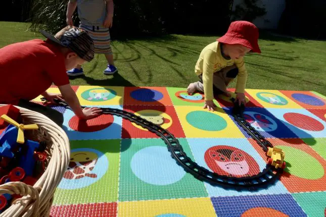 2 boys sittingon a cushioned playmat, playing with a train track. The mat has a pattern made up of circles and squares in bright colour, with some zoo animals