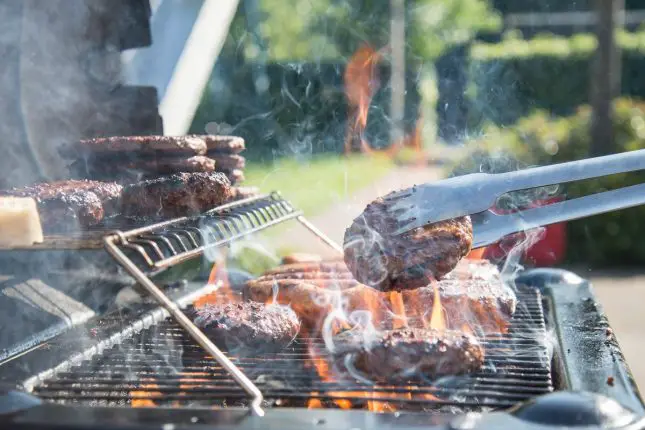A flame BBQ with burgers and sausages and a pair of tongs holding a burger