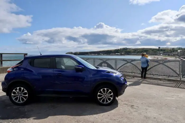 A blue Nissan Juke car parked next to the beach. A woman and child are in the background looking at the sea