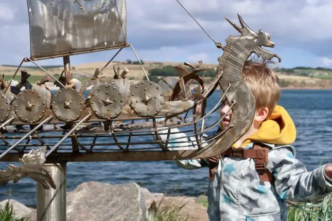 A toddler standing behind a metal boat sculpture