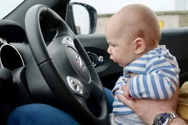 A young baby sitting on his dads knee in the drivers seat of the car, staring at the steering wheel