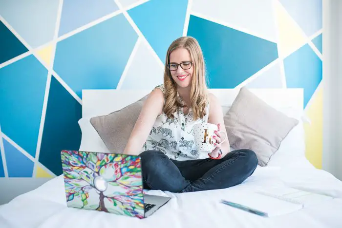 Pinterest Management Services | A blonde woman wearing glasses is sat on a bed with her legs crossed. She has on black jeans and a white shirt with zebras on it. She is holding a white mug with a gold 'K' on it and gold polka dots. She is working on an open laptop www.oddhogg.com