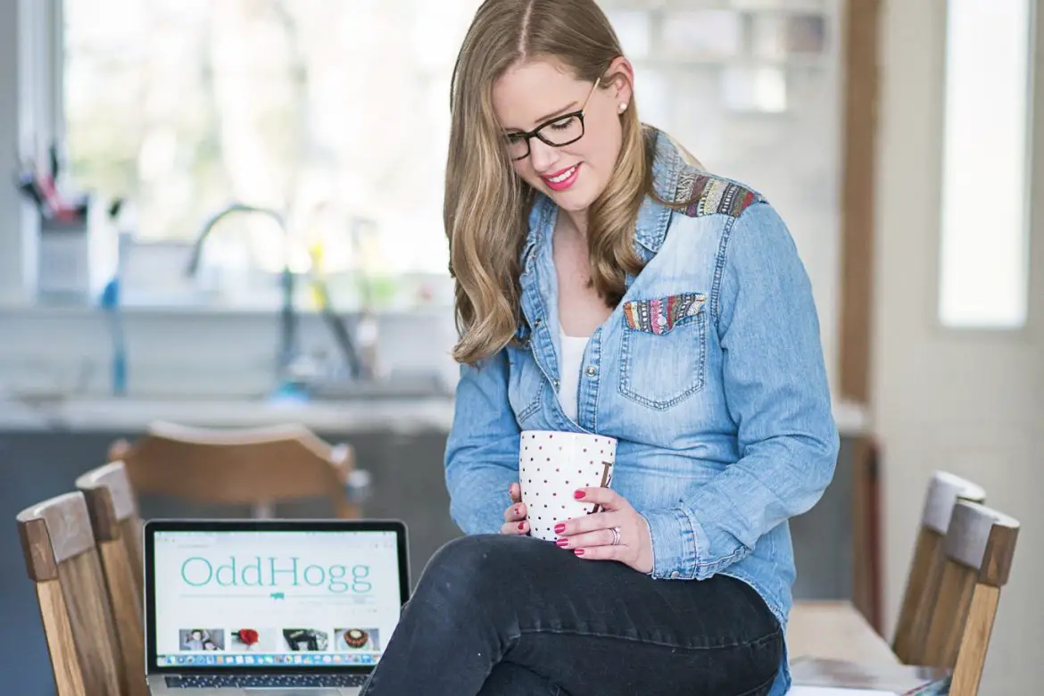 Pinterest Management Services | Woman sitting on the kitchen table wearing black jeans and a denim shirt. She is holding a white mug with gold polka dots on it. Next to her is an opten laptop www.oddhogg.com