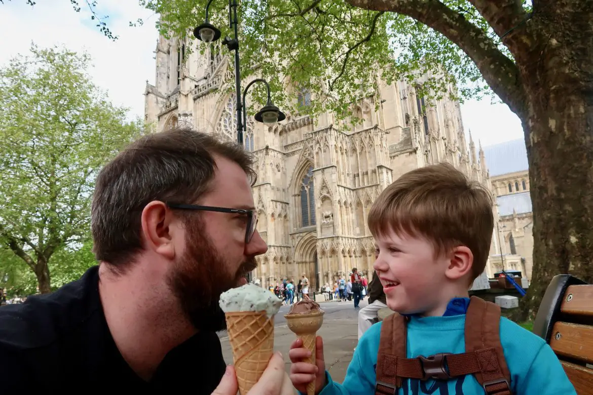 A young boy in a blue jumper is eating a chocolate ice cream with his father, eating a mint ice cream, in front of York Minister