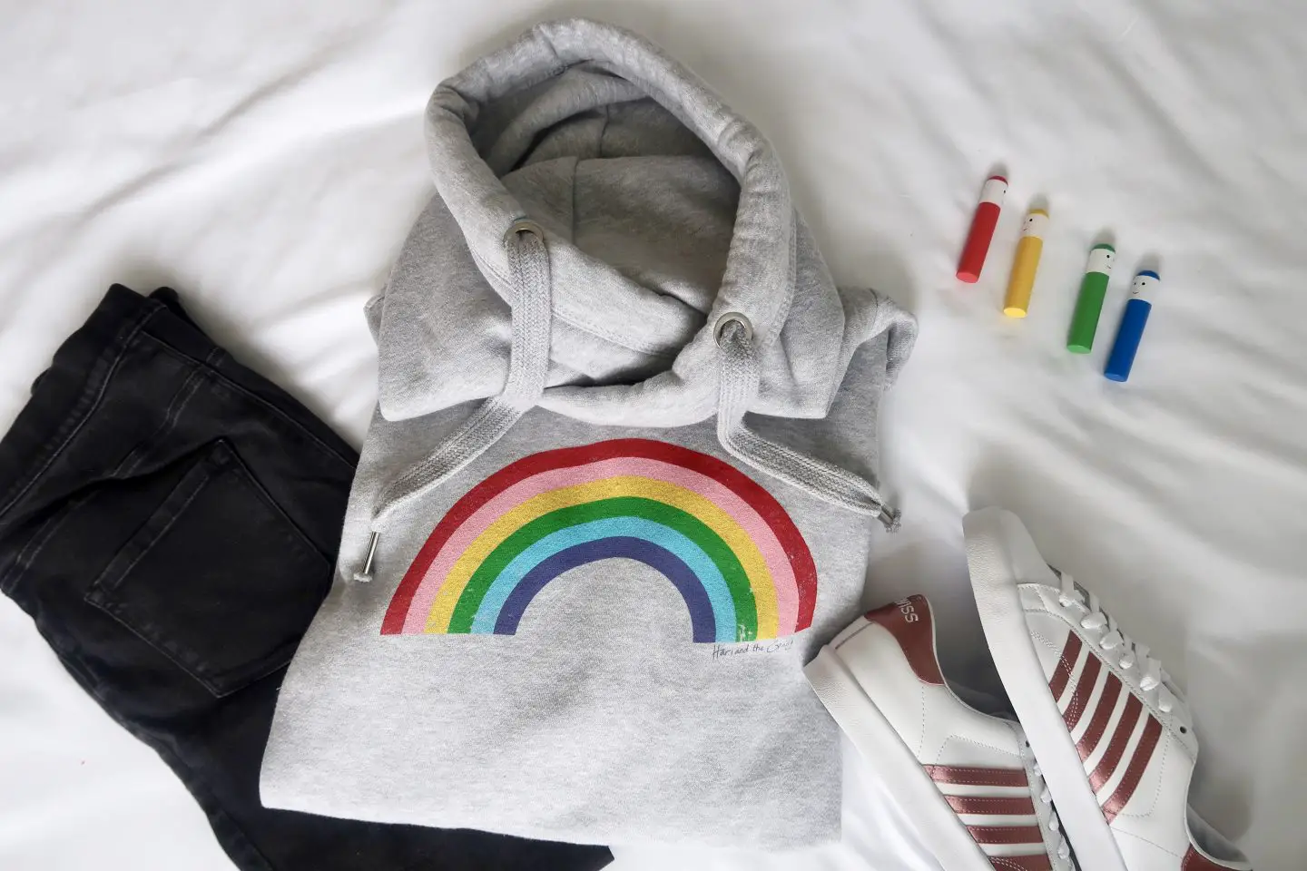 A rainbow hoodie, black jeans, white trainers and 4 rainbow coloured peg dolls lying on a bed