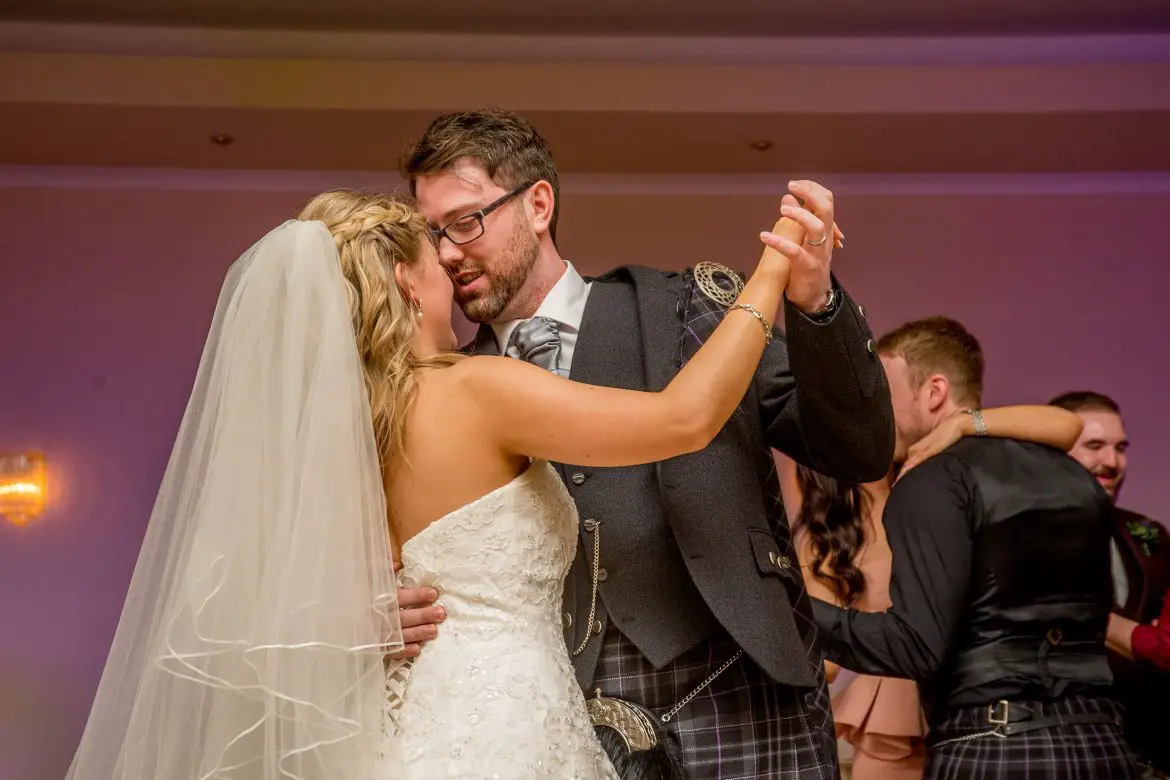 A bride and groom dancing. The bride has blonde hair and is wearing a white strapless dress and a veil. The groom is wearing a grey and purple kilt with a grey jacket.