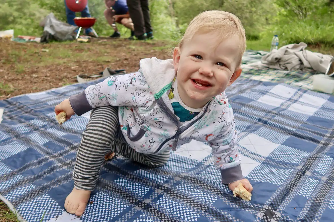 A toddler on a rug in the forrest. He is wearing stripy trousers and a white hoodie. He is smiling and has blonde hair