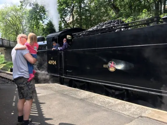 A man holding a young girl in his arms. they are watching as a black steam train goes past on a sunny day in Bradford