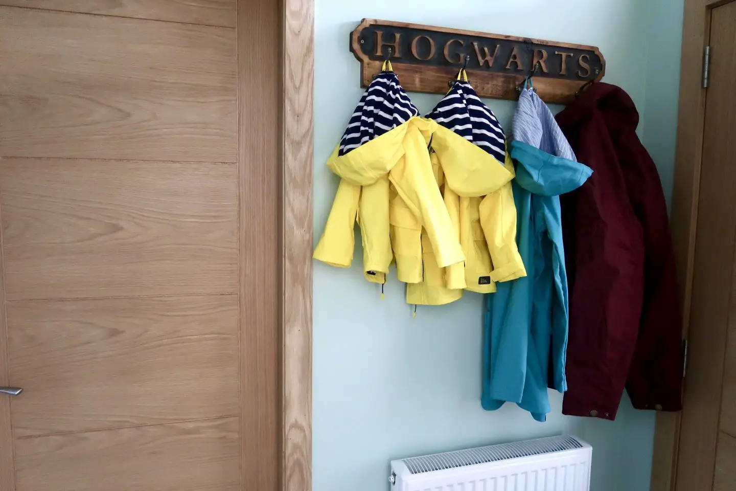 A hallway with oak doors and light green walls. There is a wooden coat hook with "Hogwarts" written on it and 4 cots hanging. There are 2 bright yellow coats with blue and white stripped hoods, one teal coat and one dark red coat.