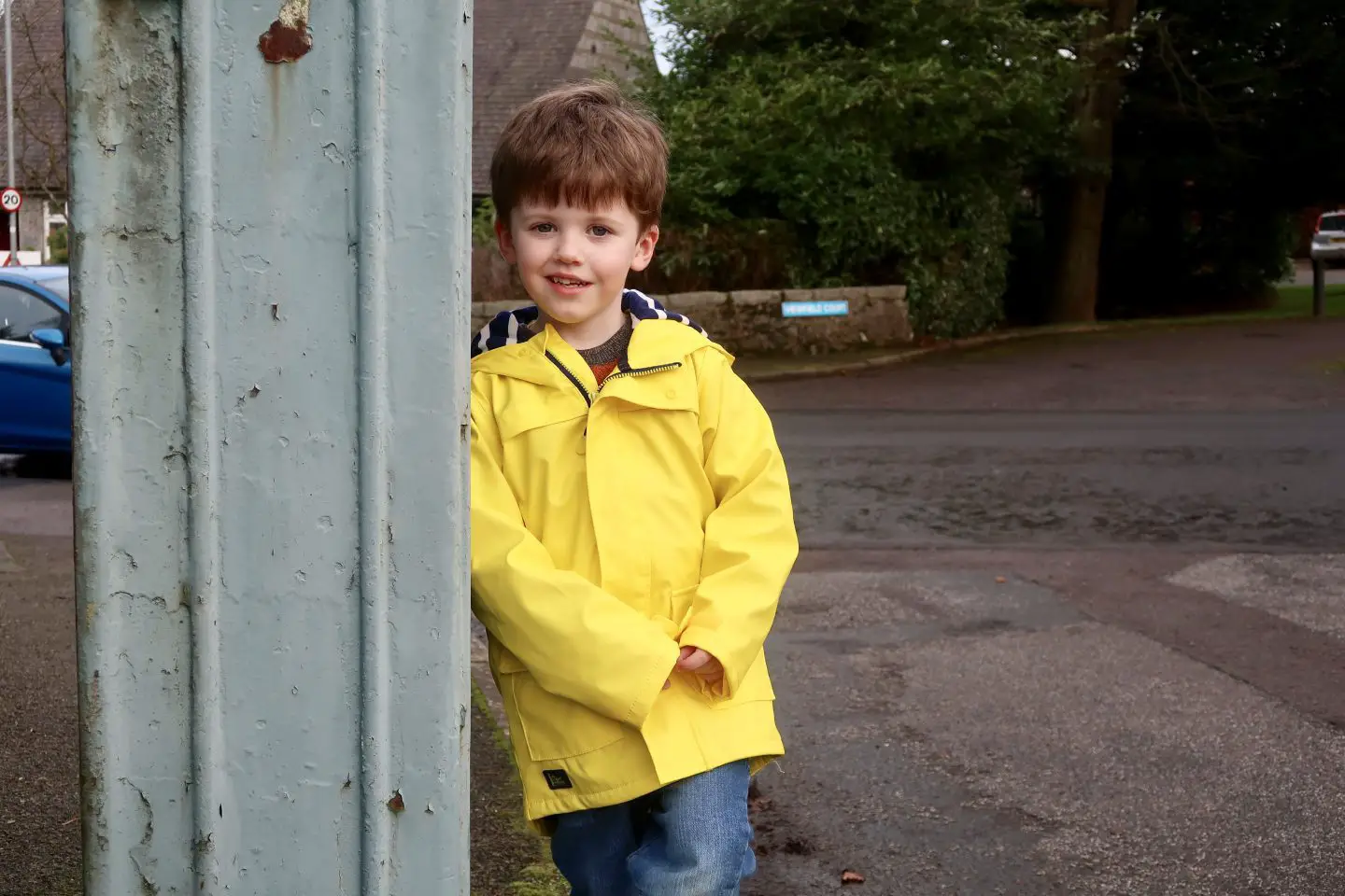 A boy in a yellow jacket standing on the side of the street and leaning against a green box.