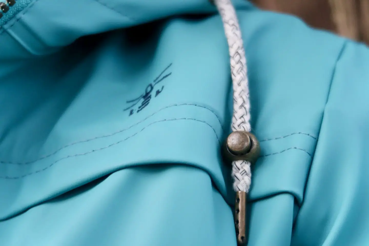 A close up of the Bowline Jacket from Lighthouse Clothing in Teal. It shows a shoulder with a cord and the Lighthouse Clothing symbol