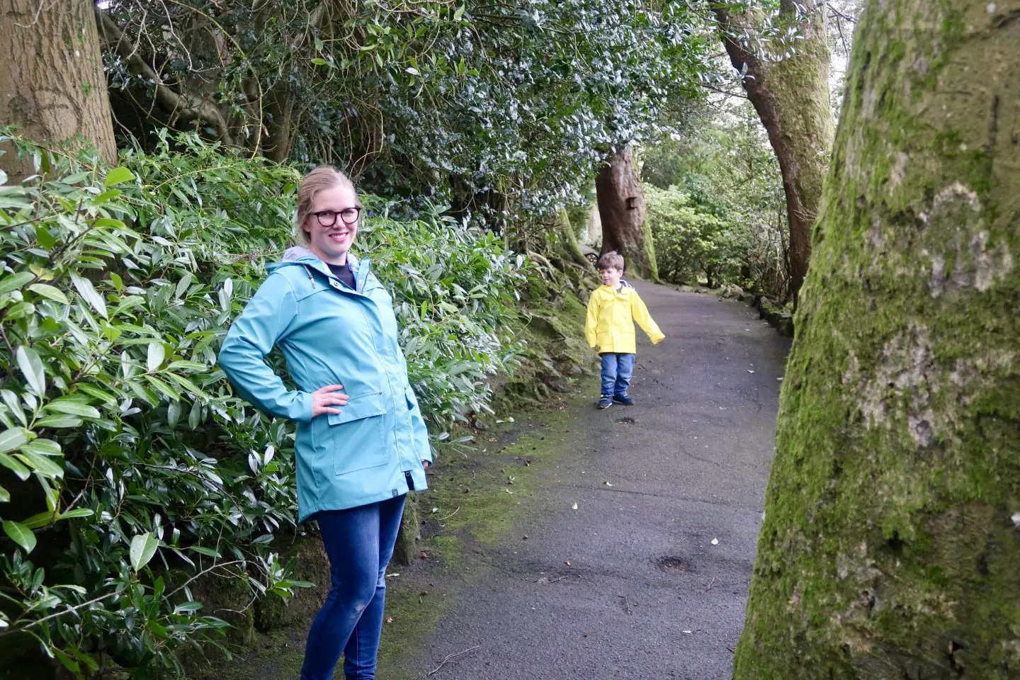 A woman standing in a teal jacket in a park, while a boy in a bright yellow jacket is in the background