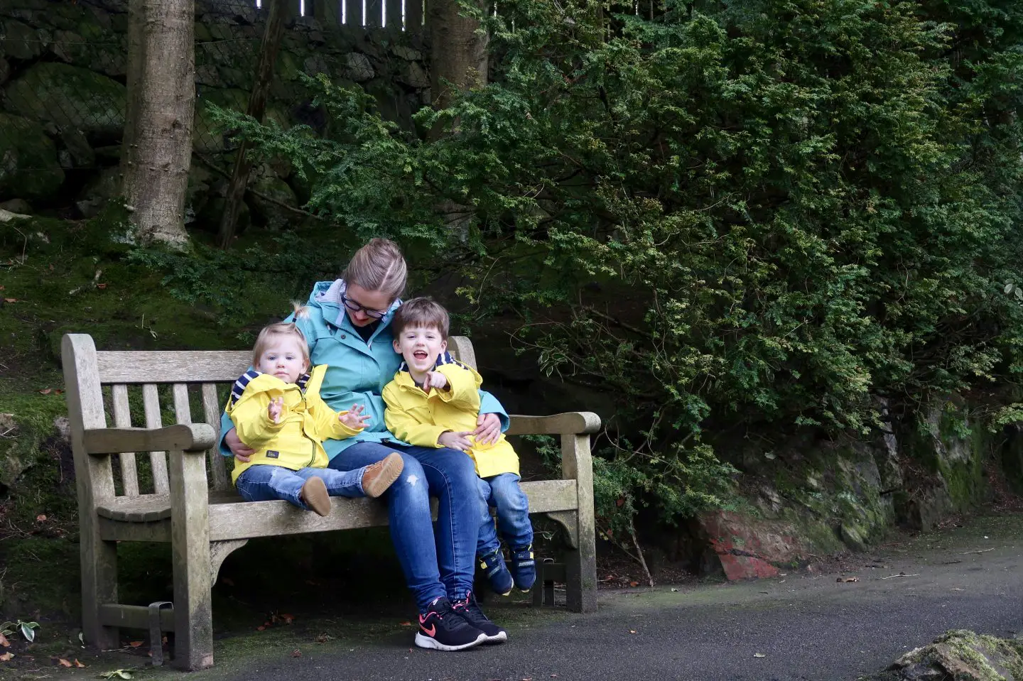 A mother and two children are on a bench in a park. The woman is wearing a teal coloured raincoat and her sons are in yellow jackets.