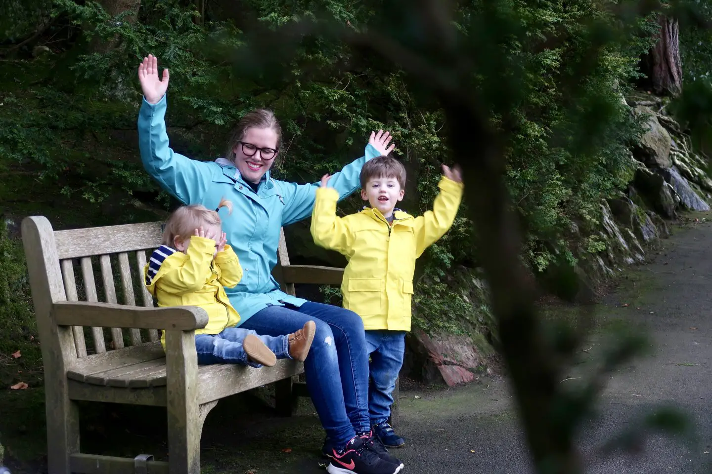 A mother and two children are on a bench in a park. The woman is wearing a teal coloured raincoat and her sons are in yellow jackets. They are throwing their hands up in celebration