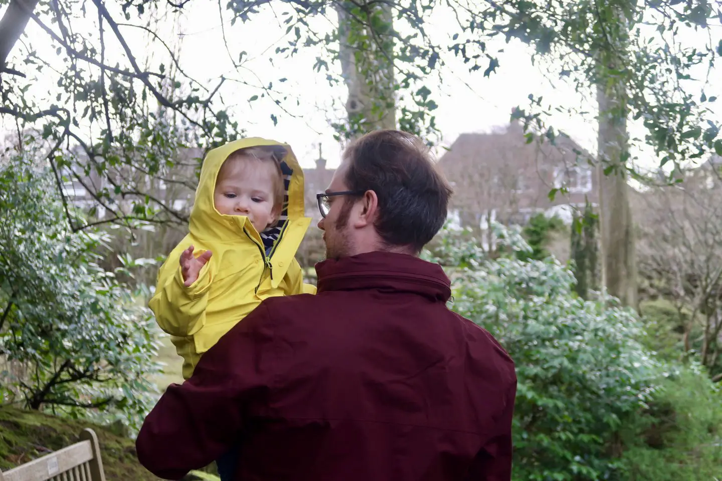 A father holding his son. He is facing away from the camera, while the boy faces the other way with the hood of his yellow jacket up