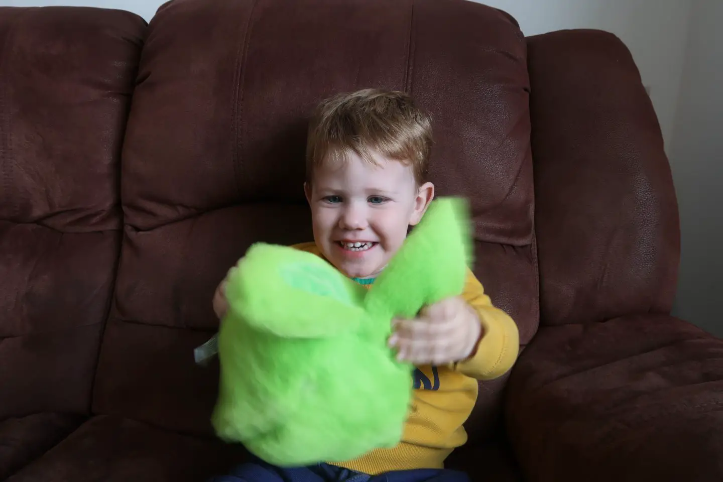 A young boy sat on a brown sofa and holding a green fluffy toy out in front of him. He is smiling at the toy