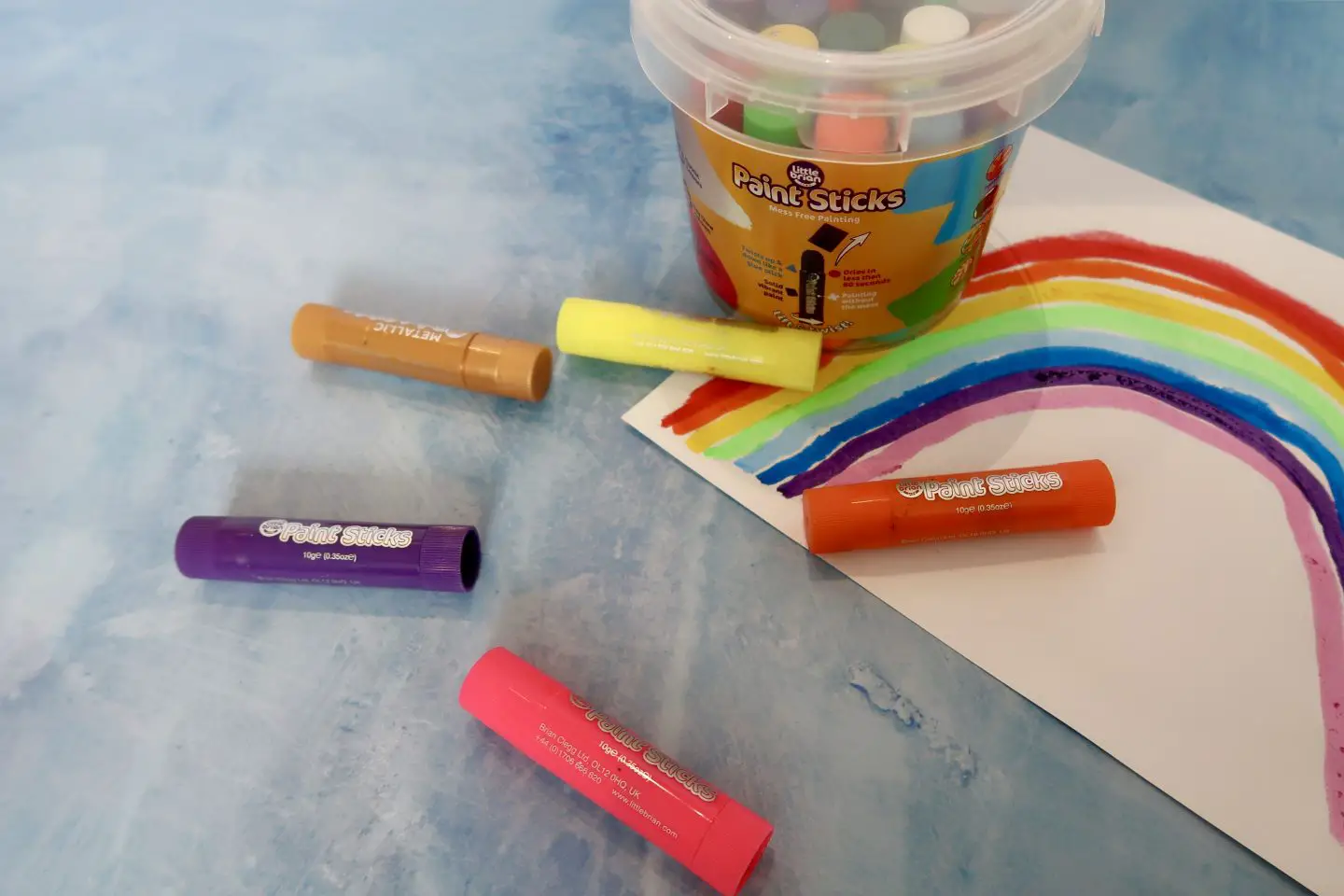 A bucket of paint sticks next to a piece of paper with a rainbow on it