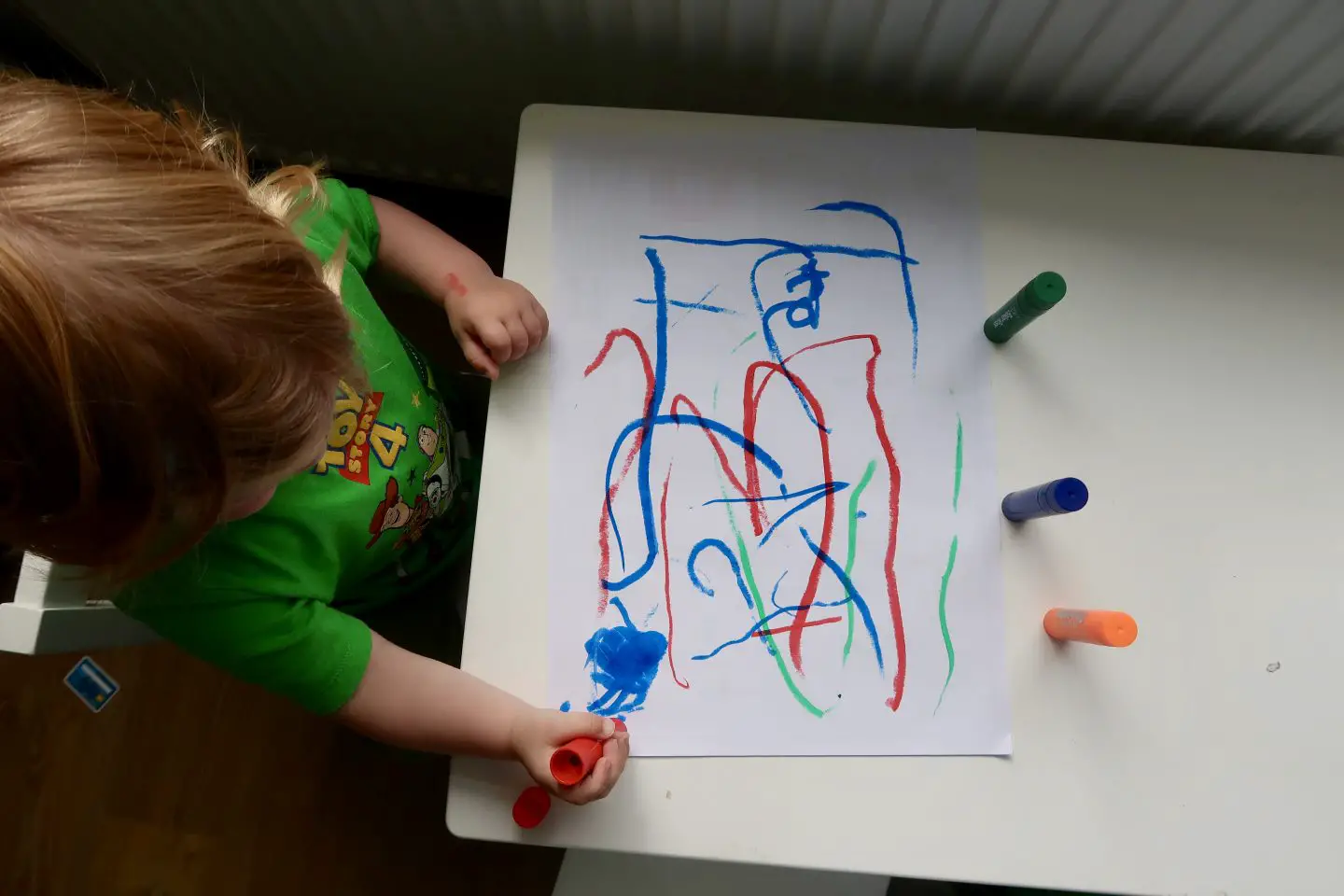 A top down view of a little boy at a small table drawing on paper with paint sticks