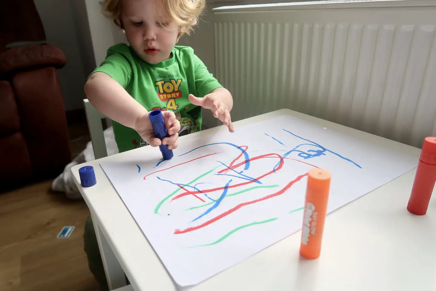 A toddler at a little table painting on paper with paint sticks