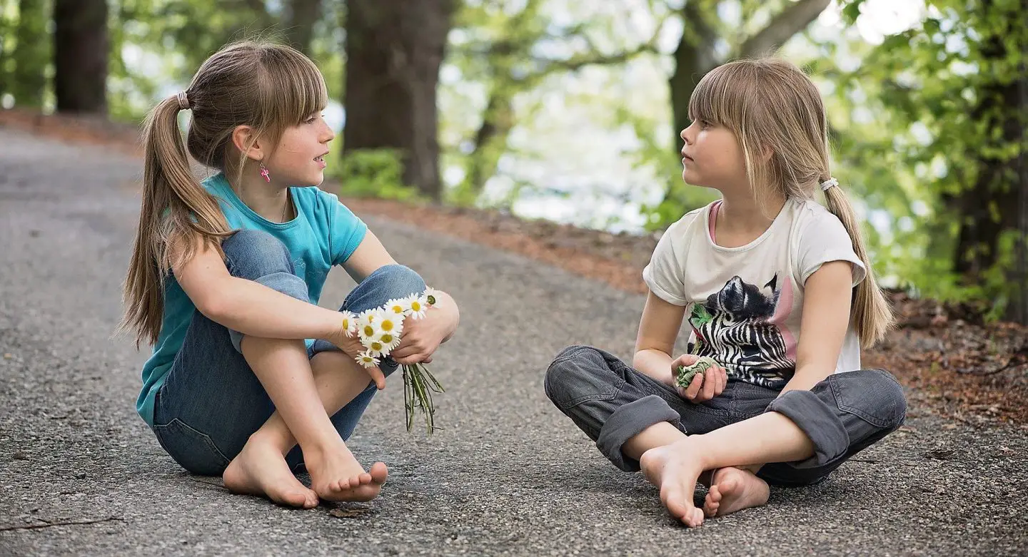 2 girls sitting cross legged on the ground on a path. The are both holding flowers and looking at each other.