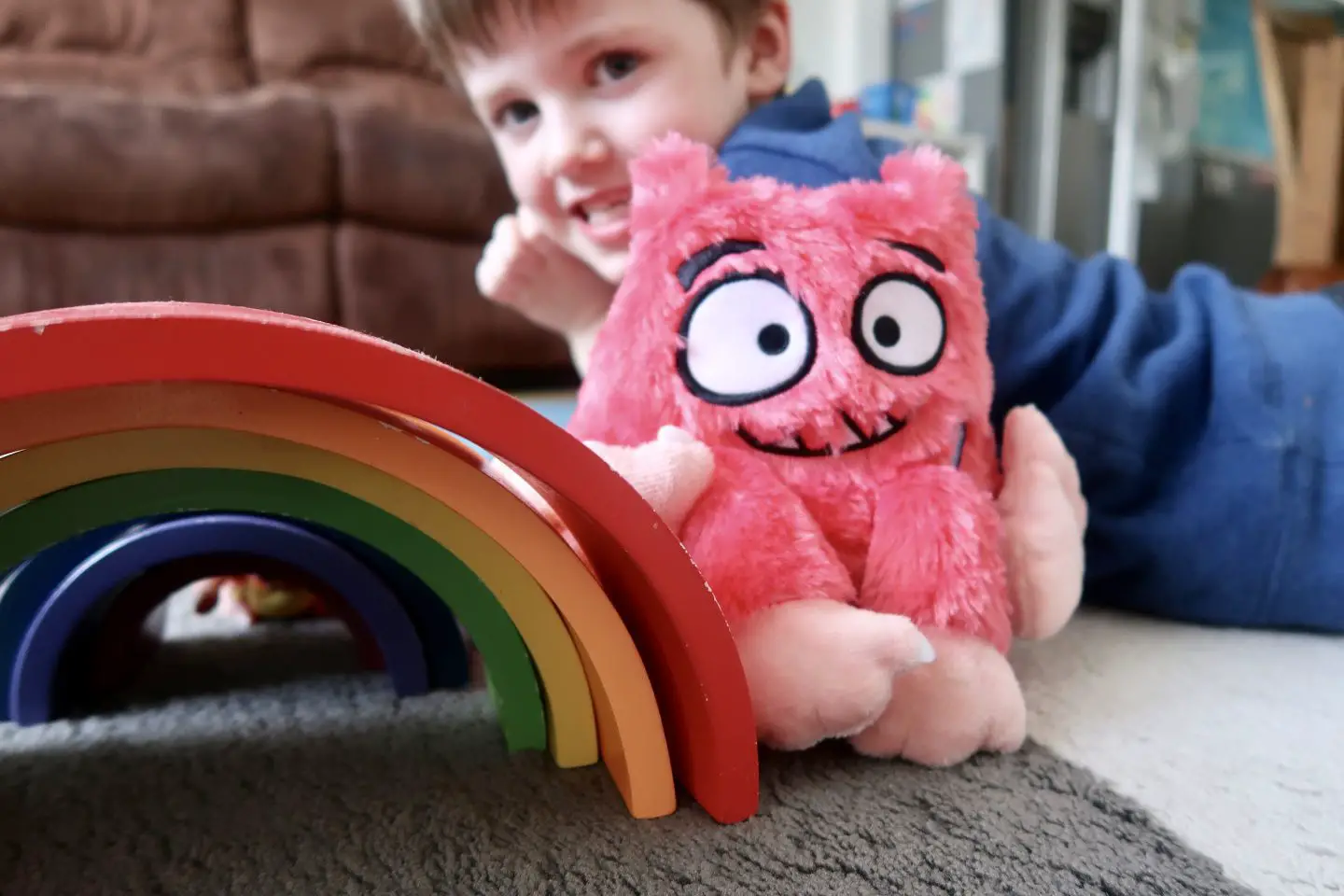 A wooden rainbow next to a fluffy monster toy. There is a boy smiling in the background slightly out of focus