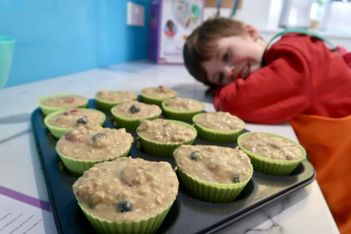 A boy resting his head on a counter and smiling at a tray of muffins about to go in to the oven