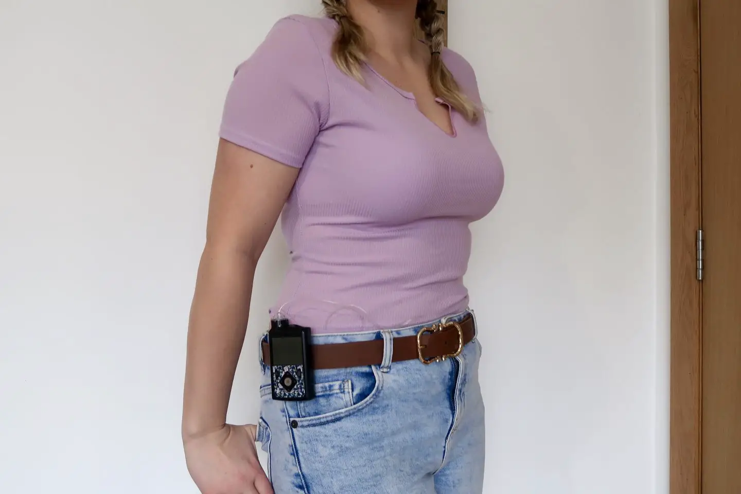 The torso of a woman wearing a lilac t-shirt and light blue jeans. She has an insulin pump on her hip and her thumbs in her back pocket.