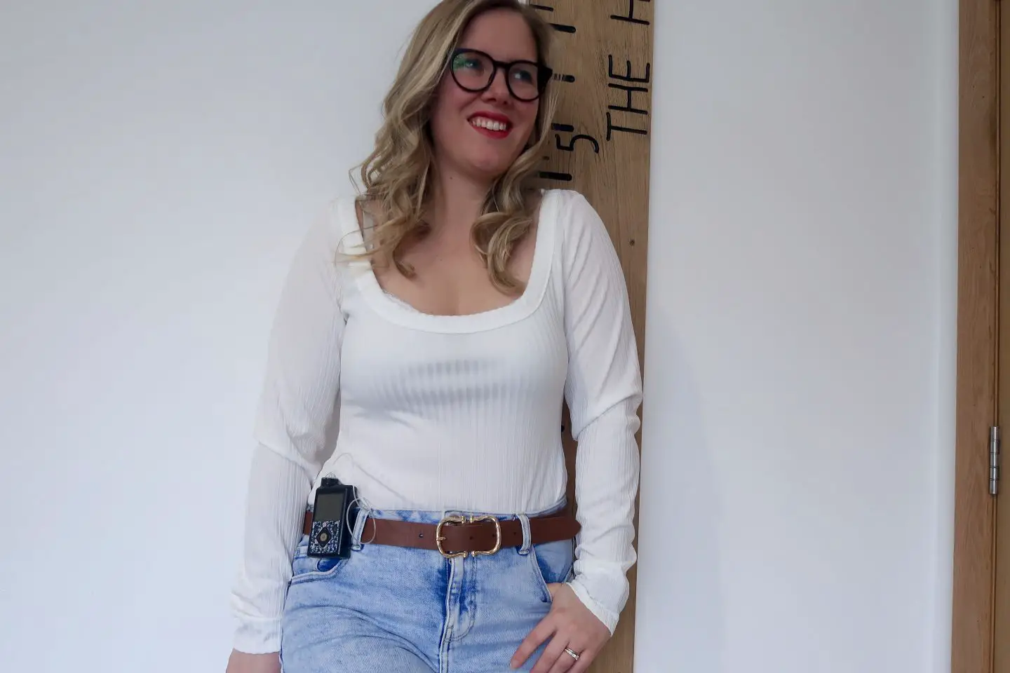 A woman wearing a cream long sleeved body suit, high waisted light denim jeans and a brown belt. She has a medtronic insulin pump clipped to her belt. She has a hand in one pocket and is smiling, looking slightly away from the camera