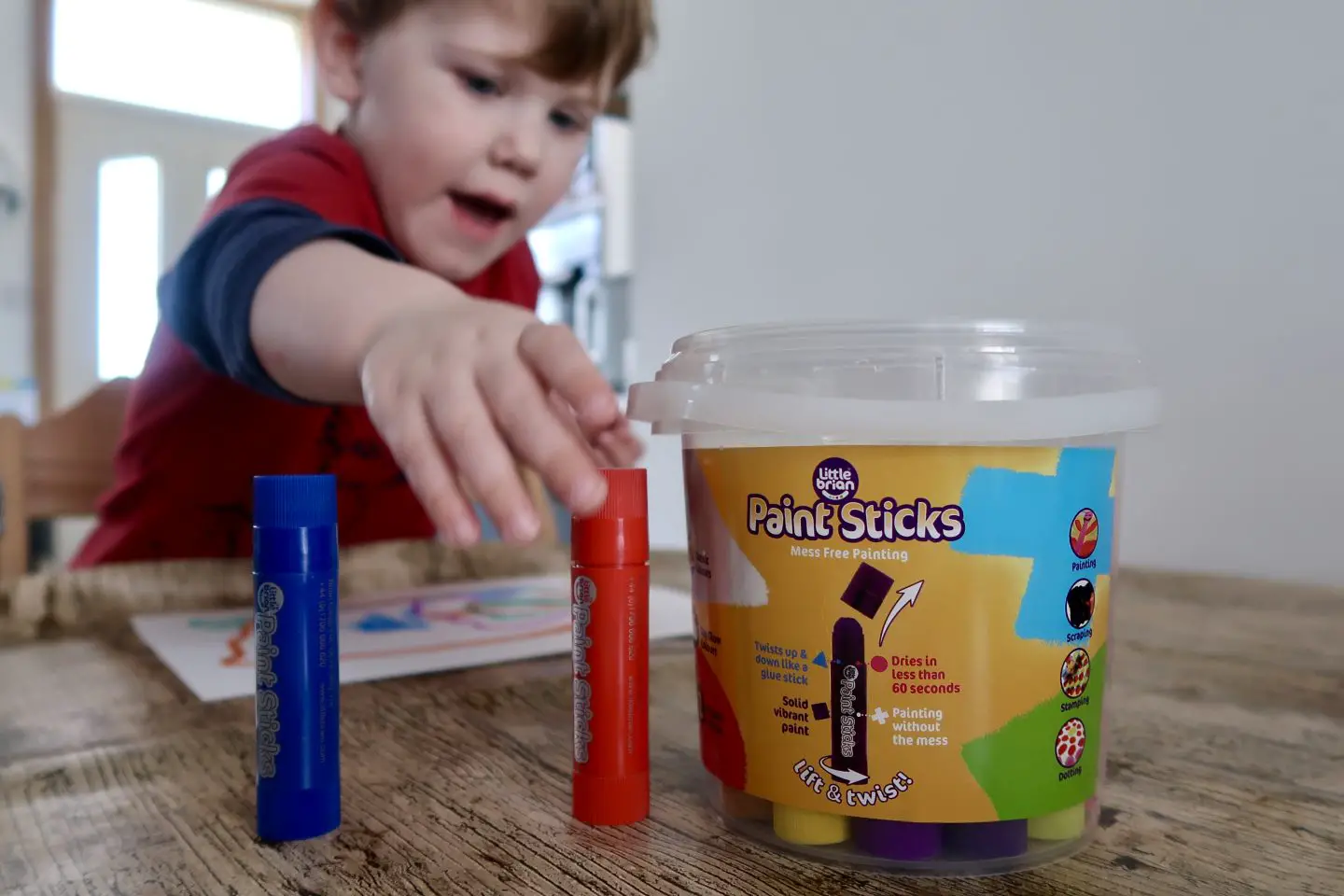 A bucket of Little Brian Paint Sticks, with a child reaching forwards to grab a paint stick
