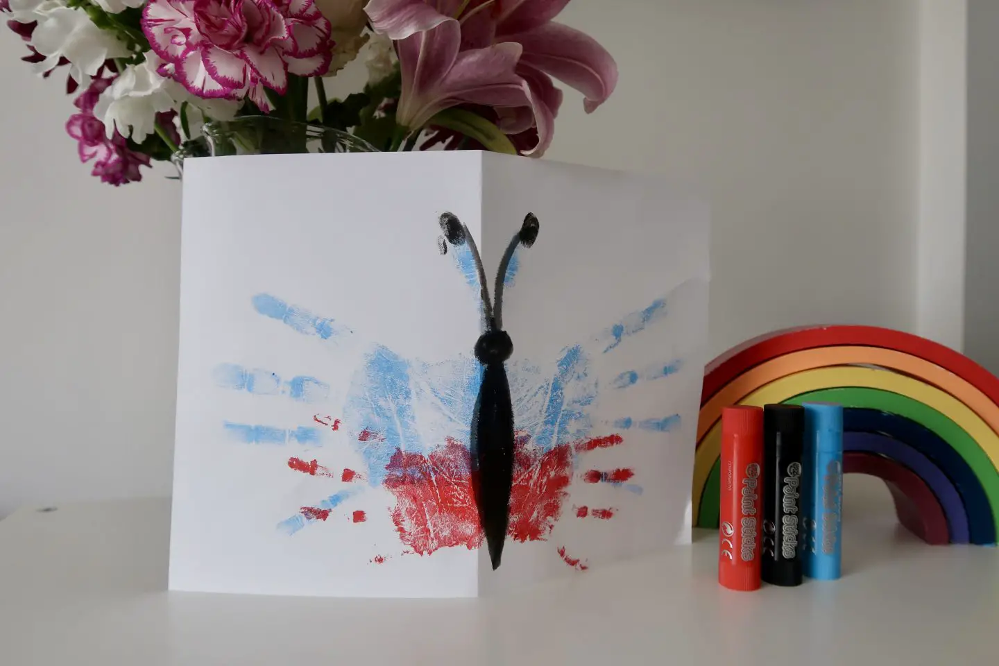 A card with a butterfly on it, made out of handprints, next to a bunch of flowers, a wooden rainbow and some paint sticks