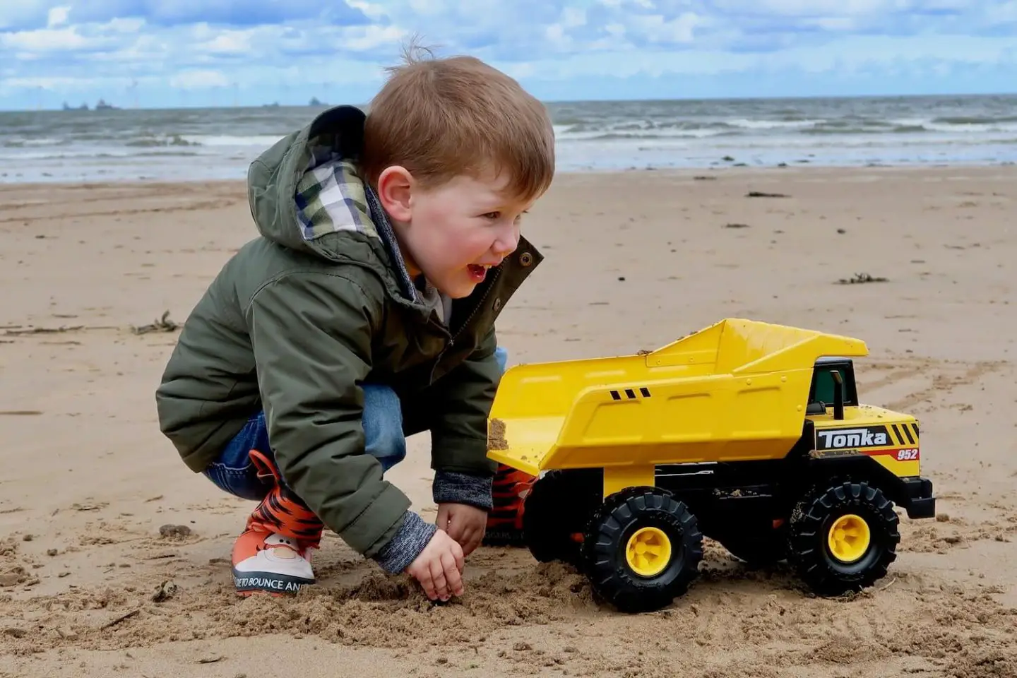 A child crouching next to a yellow toy dump truck on the beach.