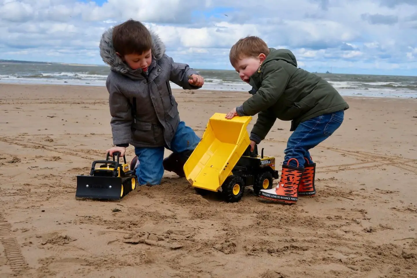 Two children play on the beach with yellow Tonka Trucks. There is a dump truck and a bulldozer. The children are animated in their play.