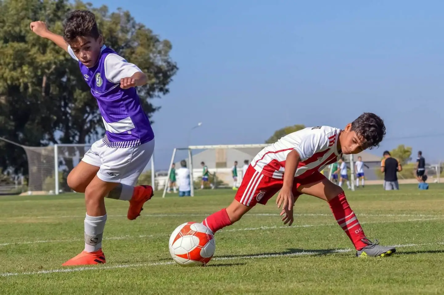 Two boys playing football. One is in a purple and white strip. The other is in a red and white strip. They are tackling each other to get the soccer ball