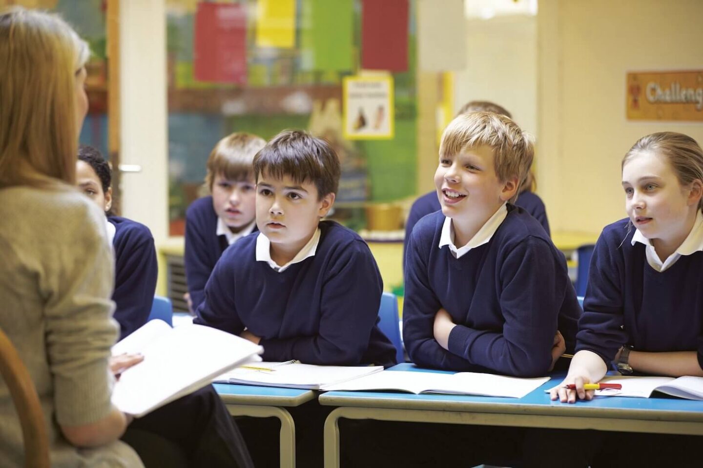 3 children in a blue school uniform sitting at a school desk looking at a teacher