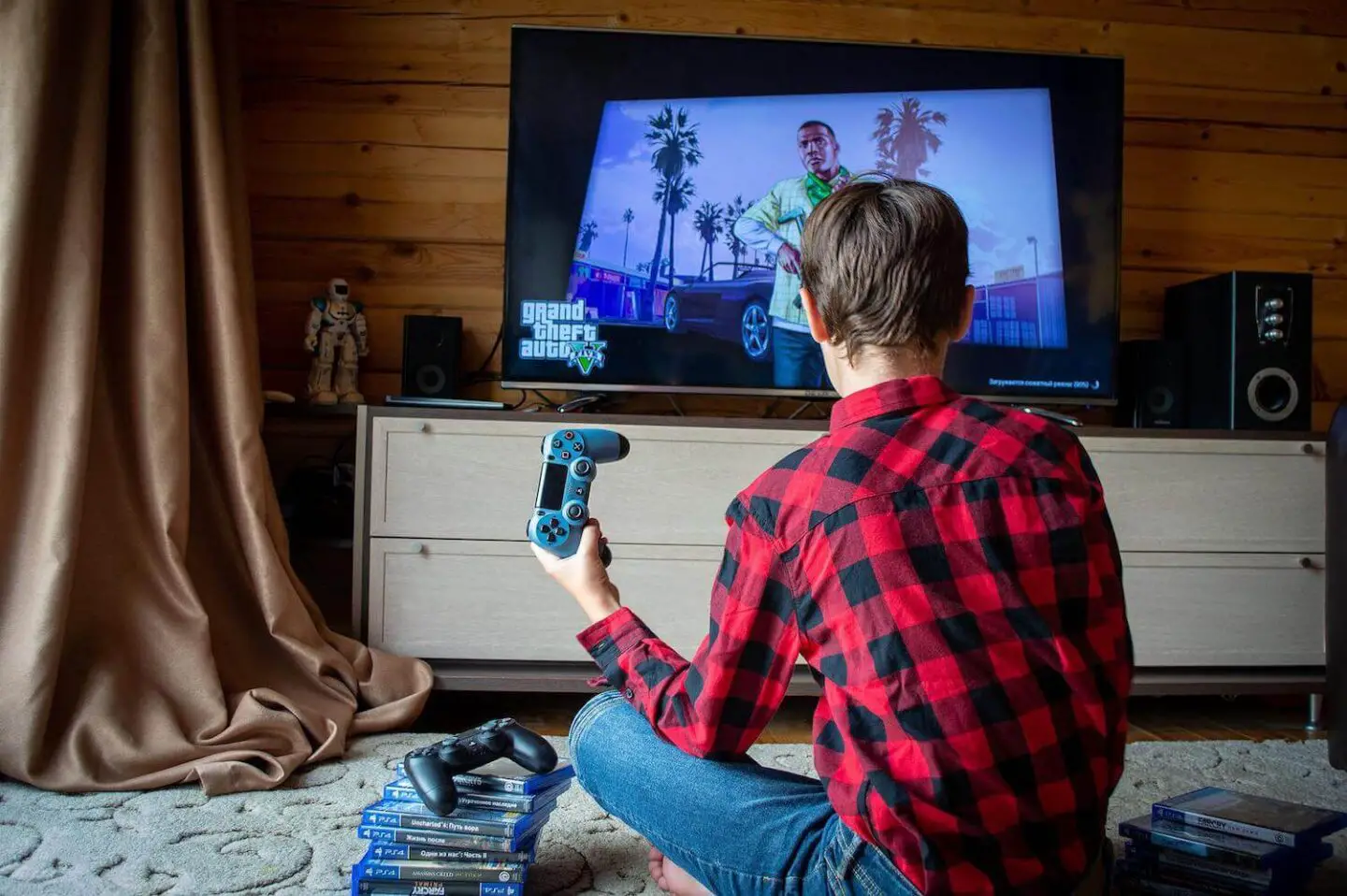 A teenage boy in a red and black checked shirt sits on the floor in front of a TV. He has holding a playstation controller and has a stack of playstation games next to him