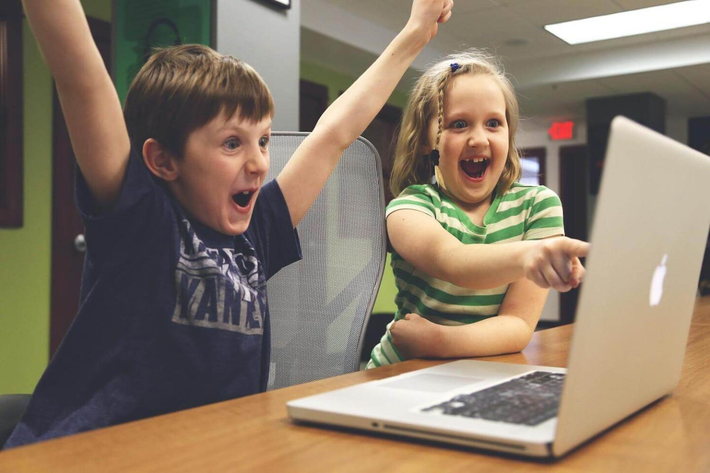 2 children sit in front of a laptop. One is cheering and the other is point at the screen with a big grin on their face
