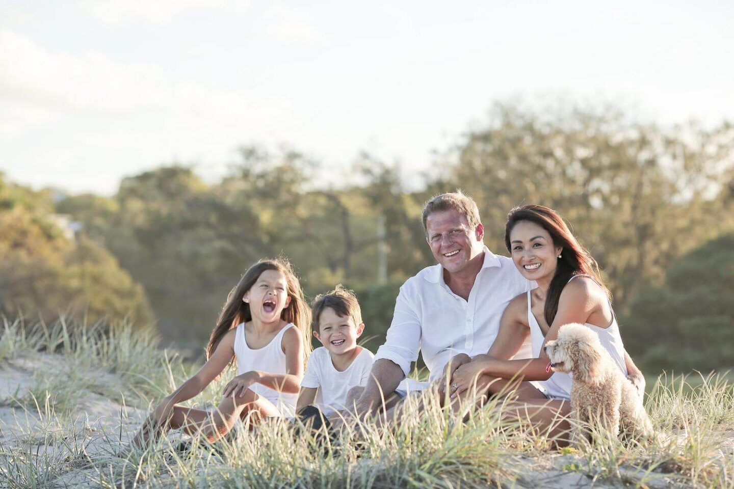 A family sit on some grass and sand with their dog. They are all dressed in white and smiling at the camera