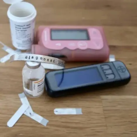 A pink Medtronic Veo insulin pump, a contour next blood glucose monitor, an insulin vial and some test strips on a table. There is also a sliver bangle.