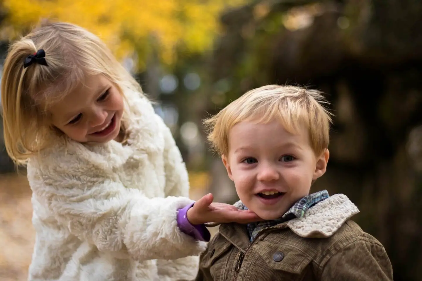 Two blonde siblings. The sister is touching the brothers chin, as if to turn his face towards her. They are wearing jackets and are outside