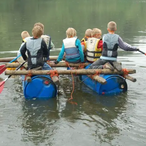 A group of school aged children rowing a homemade raft. It is made out of barrels, logs and rope
