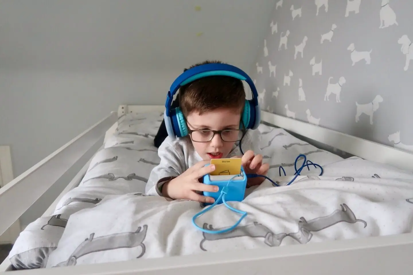 A boy lying on a bed looking down at a Yoto mini player. He is wearing blue headphones and his hand is reaching to touch the yellow card in the top