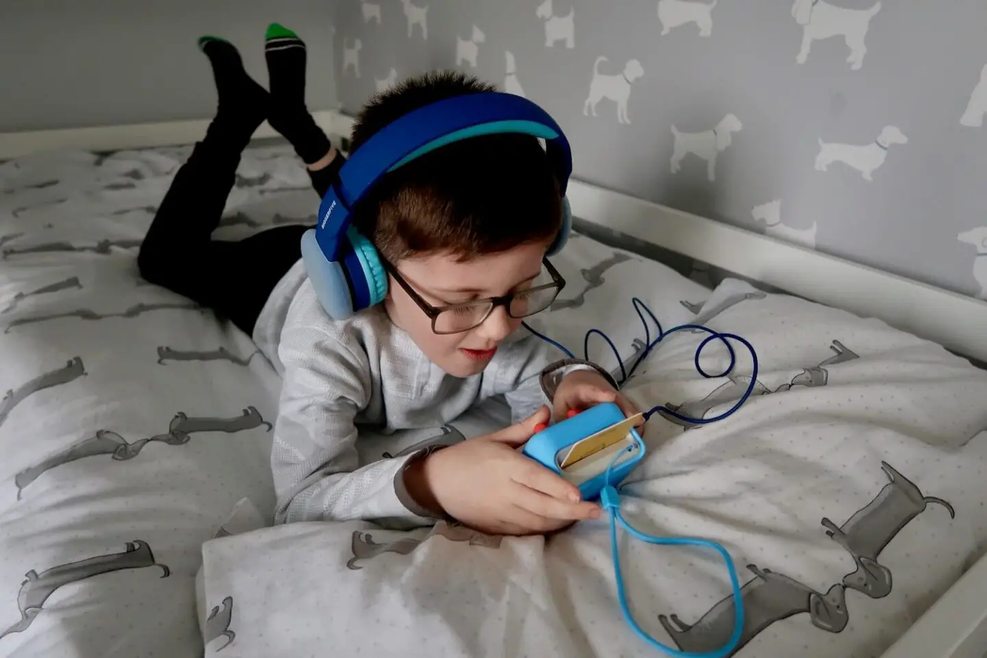 A boy lying on a bed looking down at a Yoto mini player. He is wearing blue headphones