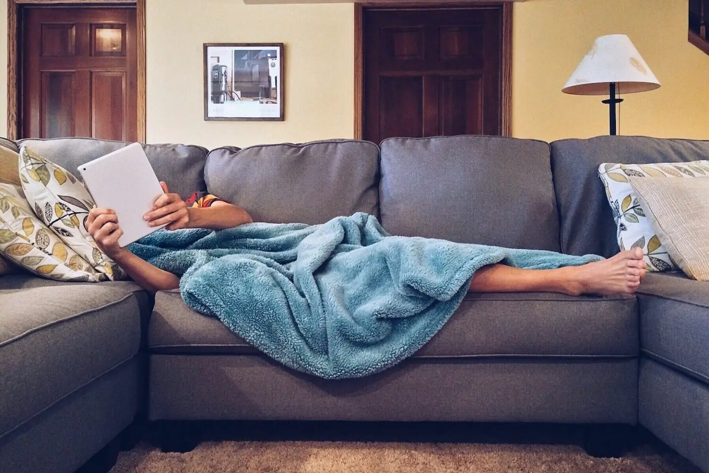 A person lying on a grey sofa. Most of their body is covered by a blue blanket and they are holding up an i-pad that covers their face.