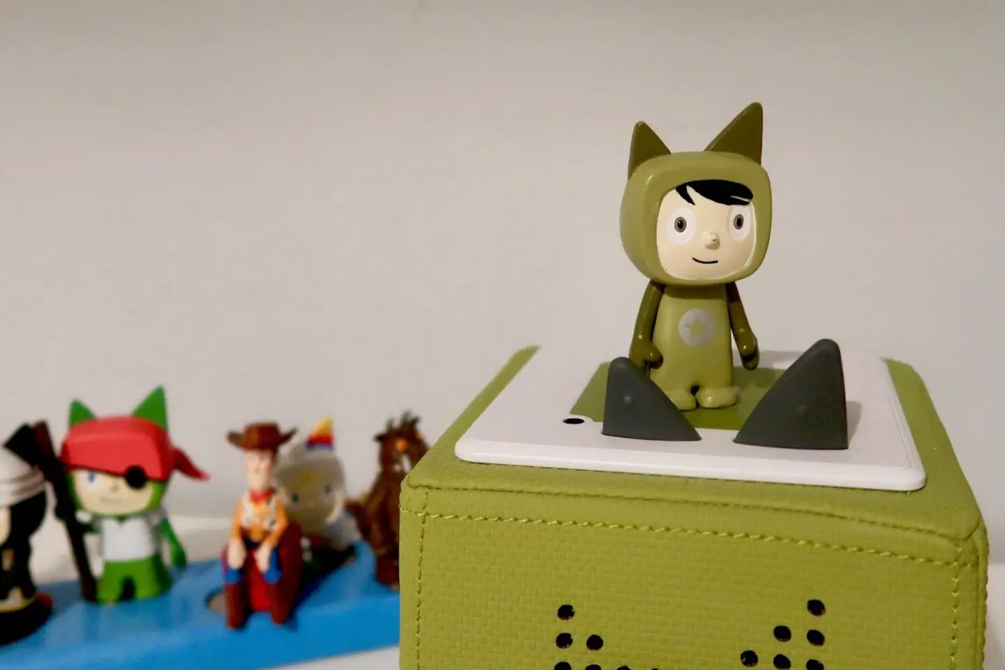 A green toniebox with a green creative tonie on the top. In the background and slightly out of focus are some other Tonies, including Woody, the Gruffalo and a pirate